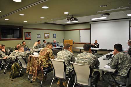 The image depicts a group of military personnel in a classroom setting. The soldiers, dressed in various military uniforms, are seated around tables arranged in a U-shape, facing a speaker at the front of the room. The speaker, also in uniform, is sitting in front of a large projection screen, leading a discussion or presentation. The room is well-lit, with overhead lights and a ceiling-mounted projector. The walls of the classroom are adorned with framed pictures and military insignia. The atmosphere suggests a formal, instructional environment, likely focused on military education or training.